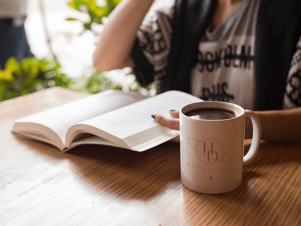 A person is reading a book in a mood lit cafe. There is a mug full of coffee with the HD Astrology logo: the letters HD within line art of Saturn.