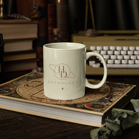 White mug on top of a book. The logo for HD astrology, a traditional astrologer, is on the cup